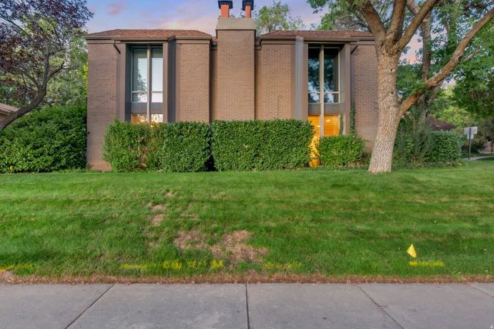 Brick exterior with large windows, mature trees, manicured shrubs, and a concrete sidewalk