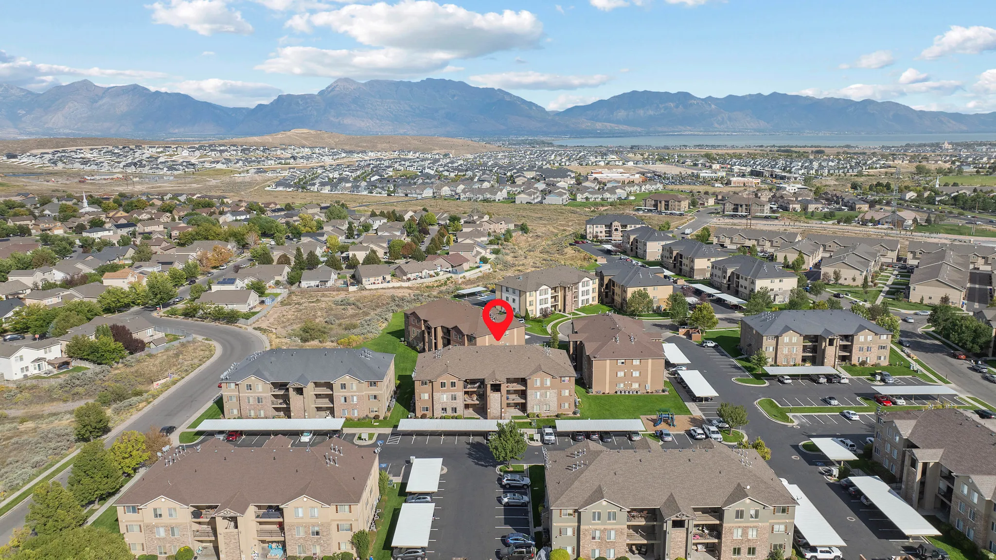 Community with multiple multi-story buildings, covered parking structures, and paved roadways, set against a mountain and lake backdrop