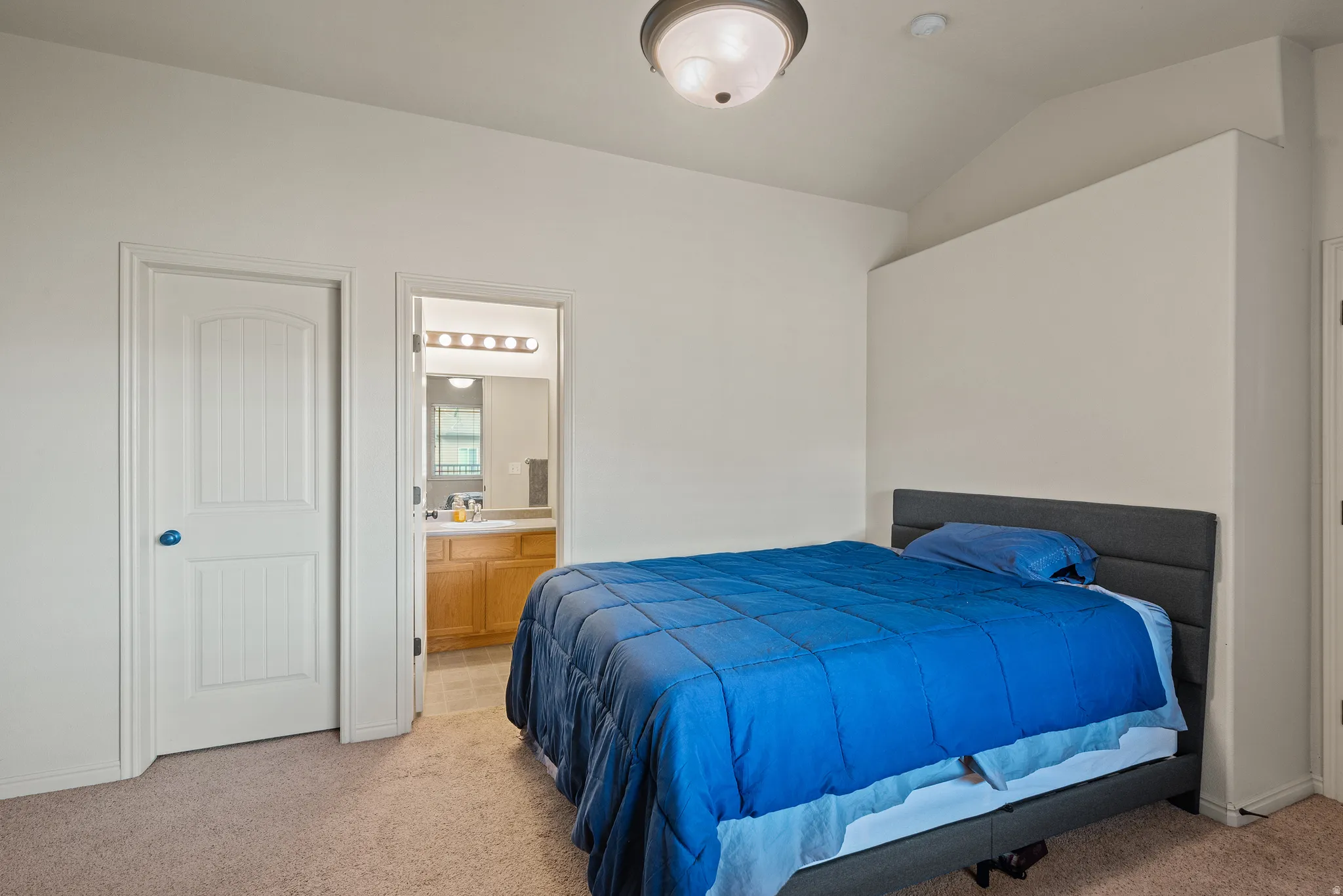 Vaulted ceiling, ceiling-mounted light fixture, light neutral carpet, paneled interior door, and an ensuite bathroom with a wood-finish vanity
