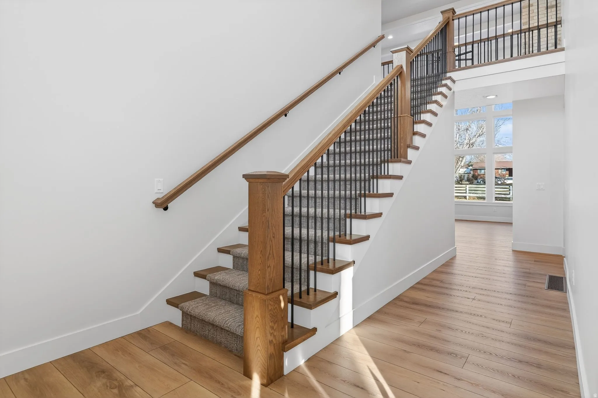 Hardwood-finish flooring, staircase with wood newel posts and handrails, wrought iron balusters, carpeted stair treads, and a multi-pane window