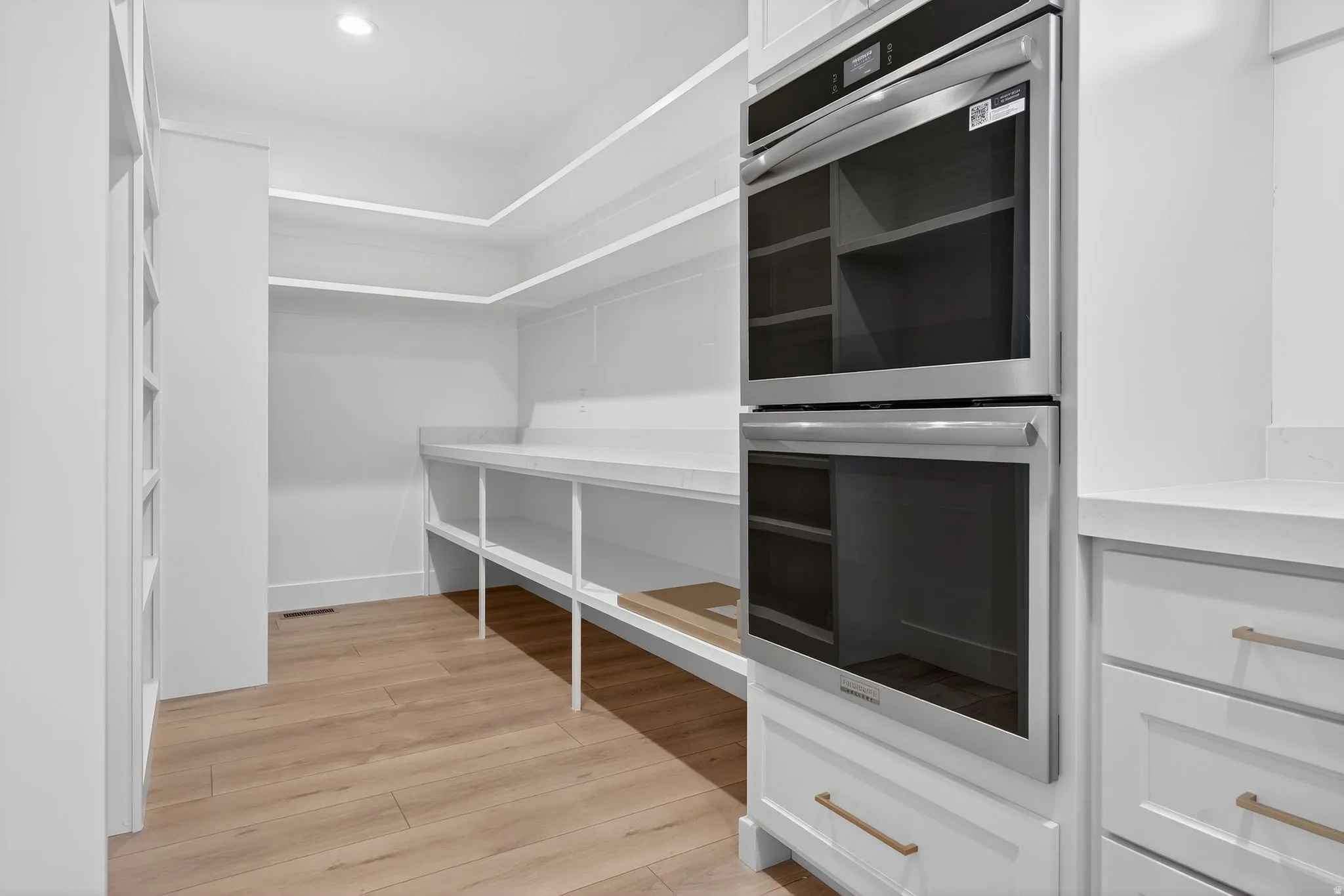 Integrated stainless steel double ovens, custom white cabinetry, light wood-finish flooring, recessed lighting, and extensive white shelving