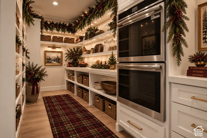 Built-in double oven, white cabinetry with gold-tone hardware, wood-finish flooring, recessed lighting, and integrated shelving