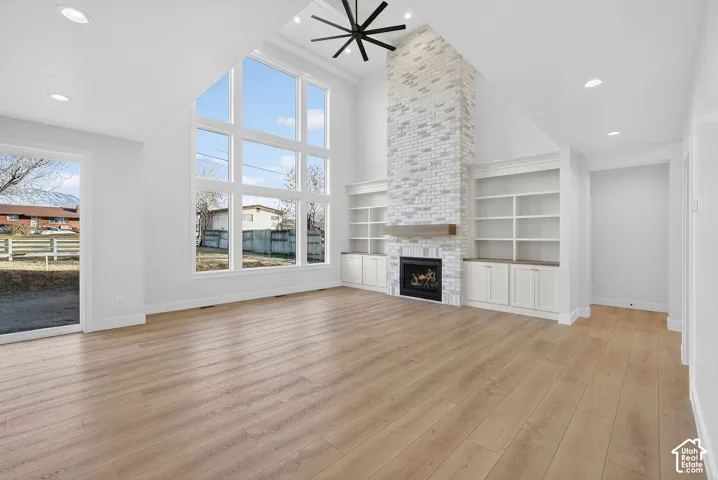 Vaulted ceilings with a ceiling fan, a fireplace with a light-colored brick surround and wood mantle, built-in shelving and cabinetry, expansive windows, and wood-finish flooring