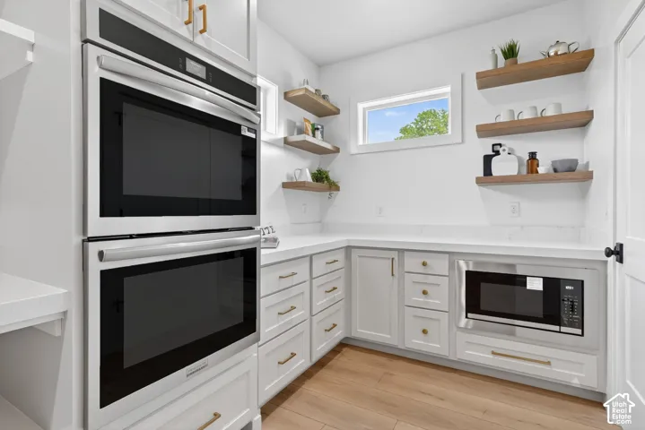 Built-in double ovens, white cabinetry with gold-tone hardware, white countertops, wood-finish flooring, and open wood shelving