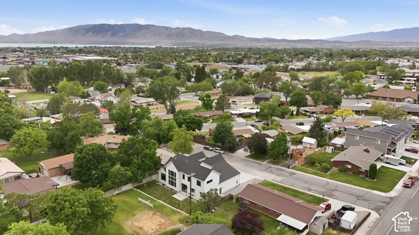 Modern exterior with white siding and dark roof, prominent two-story window array, attached garage, surrounded by mature trees and residential structures, with distant mountains and a body of water