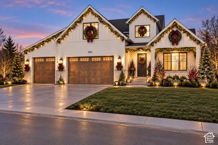 White board-and-batten siding with stone accents, dark-framed windows, wood-finish garage doors, and landscape lighting