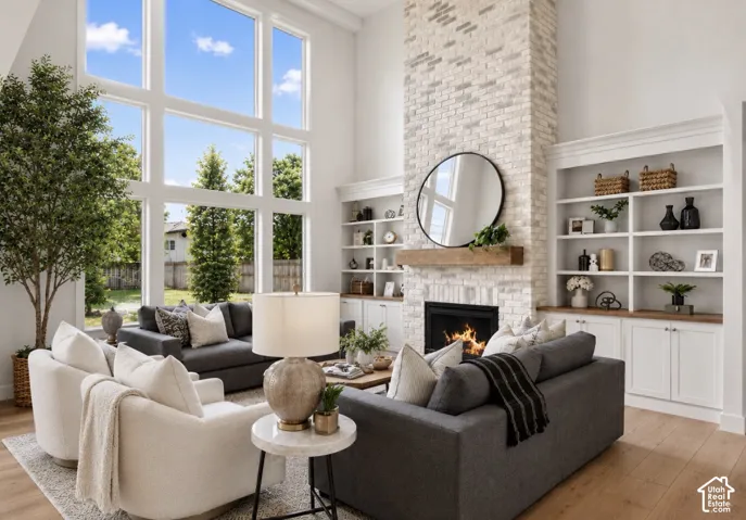 High ceilings with floor-to-ceiling windows, light wood-finish flooring, a white brick fireplace with a wood mantle, and built-in shelving with lower cabinetry