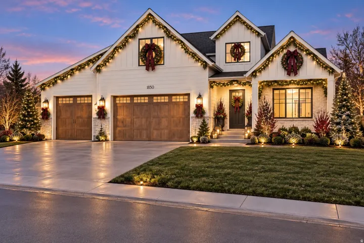 White board-and-batten siding with stone accents, dark-framed windows, wood-finish garage doors, and landscape lighting