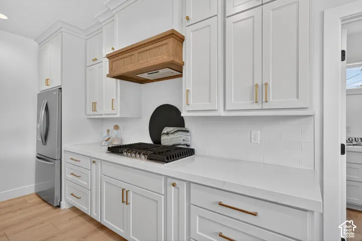 White shaker cabinetry with gold-tone hardware, wood-finish range hood, integrated gas cooktop, white subway tile backsplash, and light wood-finish flooring