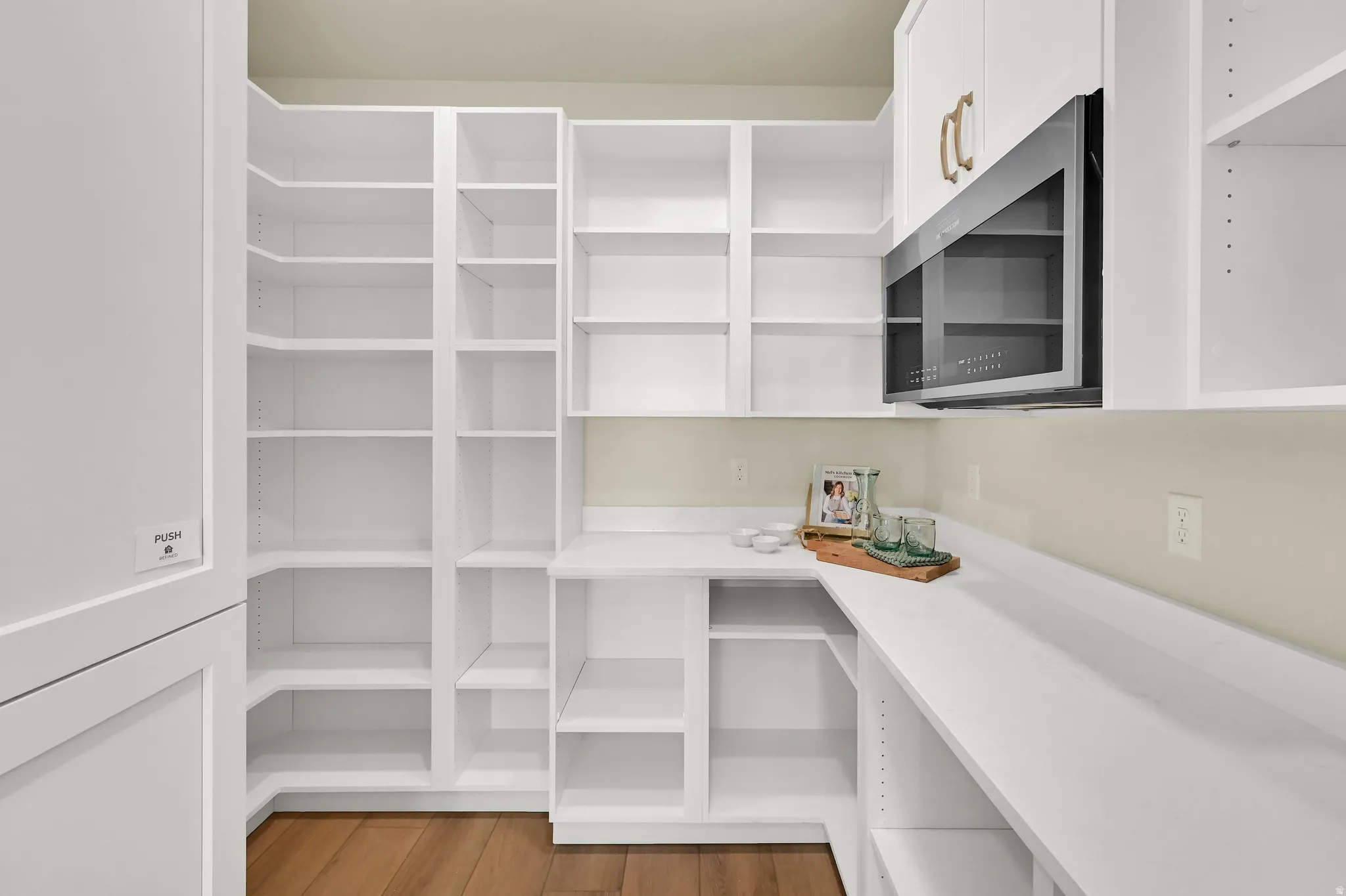 Extensive white built-in shelving, white cabinetry with gold-toned hardware, light-toned countertops, and wood-finish flooring