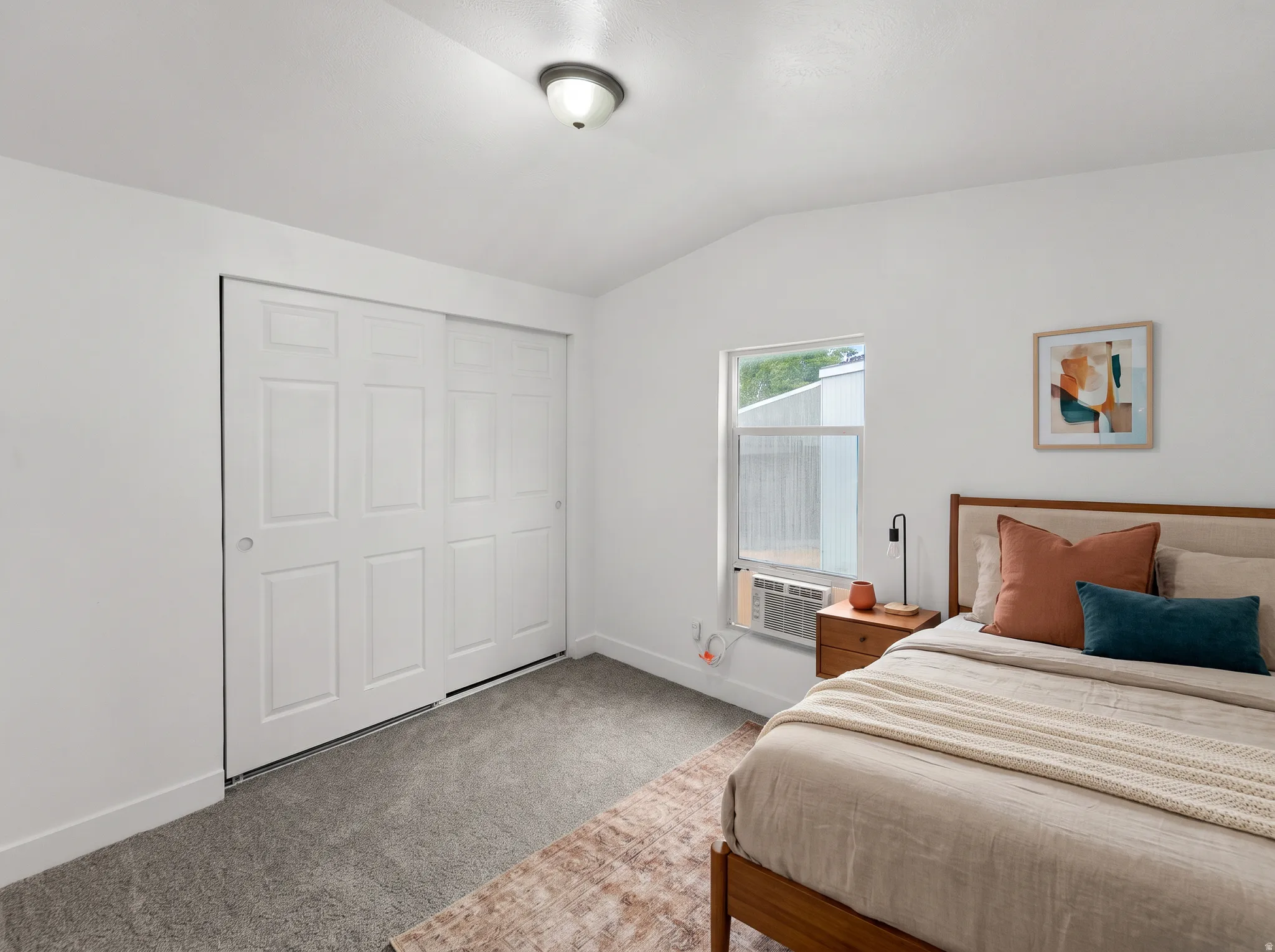 Vaulted ceiling with flush-mount lighting, sliding closet doors, carpeted flooring, and a window with an integrated air conditioning unit