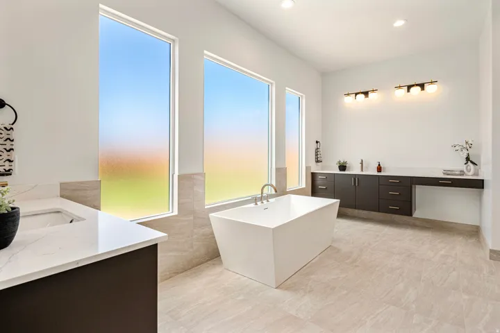 Contemporary white soaking tub, large format tile flooring, dual vanity with undermount sinks, white countertops, and expansive windows