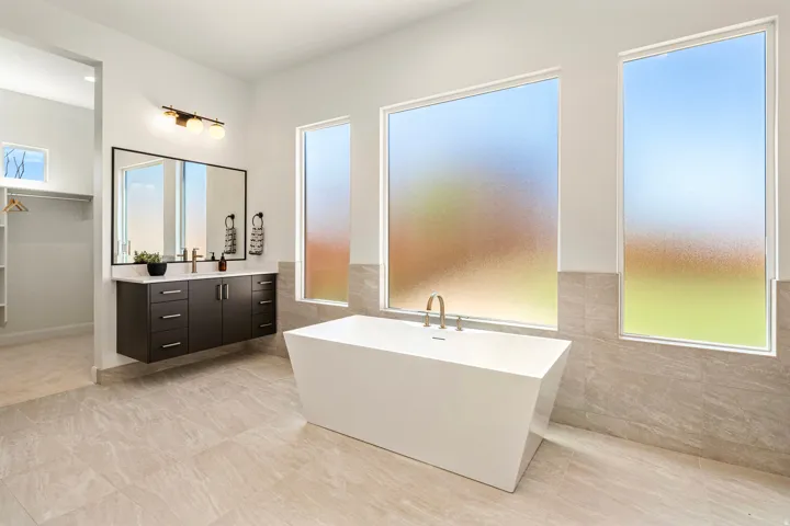 Floating vanity with dual sinks, a large framed mirror, a freestanding soaking tub, and tile flooring
