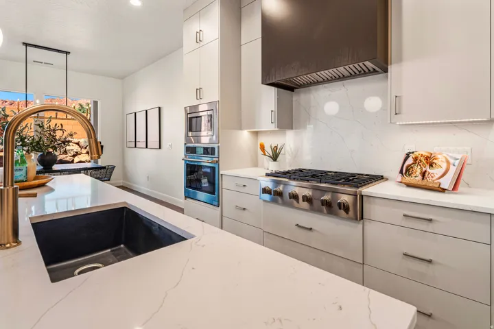 Kitchen with light gray cabinetry, built-in stainless steel appliances, a gas range with a dark range hood, a kitchen island with an undermount sink, and a gold-finish faucet