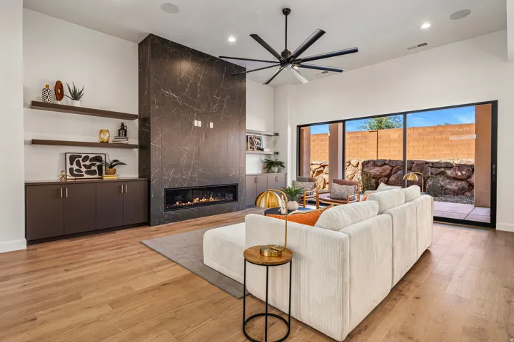 Living area featuring wood-finish flooring, a linear fireplace with a dark stone-clad surround, built-in cabinetry with open shelving, a ceiling fan, and sliding glass doors to an outdoor patio
