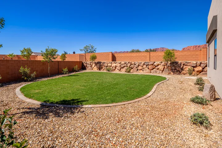 Landscaped backyard featuring a grass lawn with concrete curbing, gravel ground cover, and a rock retaining wall