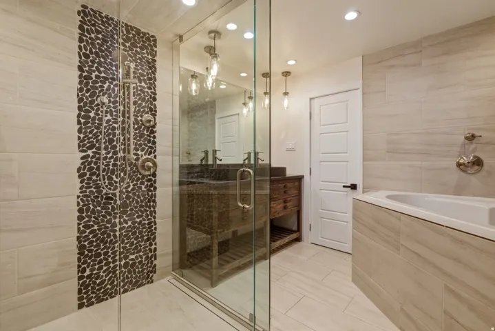 Glass-enclosed shower with pebble tile accent wall, wood-finish vanity with dark countertop, and built-in tub with coordinating tile surround