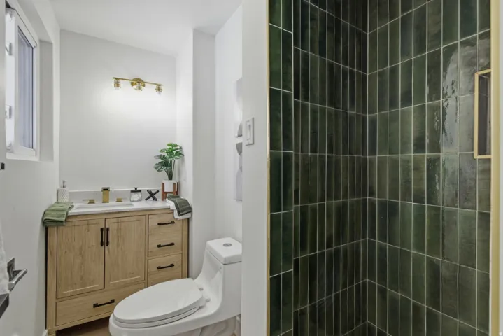 Bathroom featuring a light wood-finish vanity with a white countertop, a white toilet, and green vertical subway tile in the shower
