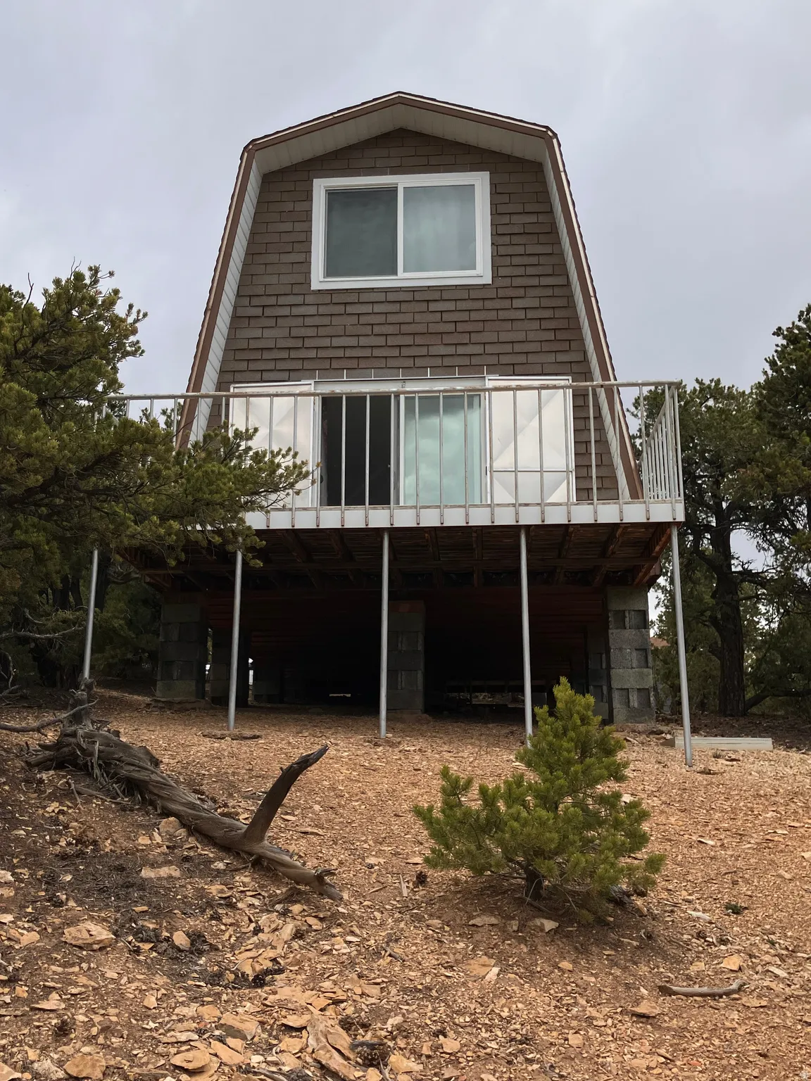 Gambrel roofline with shingle siding, elevated structure on concrete block piers, upper-level balcony with white railings, sliding glass door access, and a single-pane window