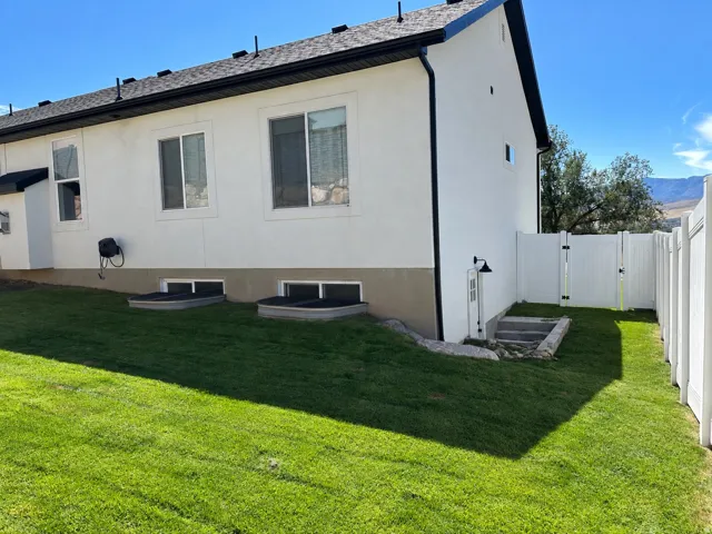 Expansive lawn, white vinyl fencing, side access gate, exterior wall lighting, and basement window wells