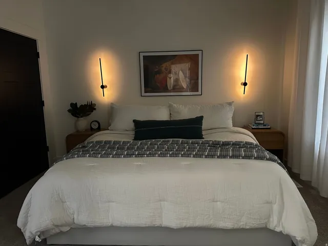 Wall-mounted sconces illuminate the space, complemented by a solid panel door, wood-finish nightstands, and light-filtering window treatments