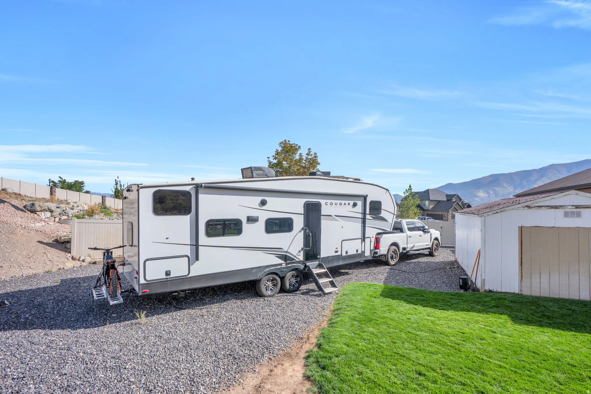 Gravel parking area, grassy yard, white storage shed, mountain backdrop, and a perimeter fence