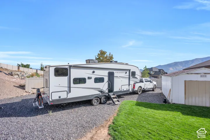 Gravel parking area, grassy yard, white storage shed, mountain backdrop, and a perimeter fence