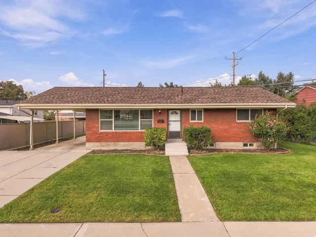 Red brick exterior, brown shingle roof, concrete driveway, carport, and a front lawn with established landscaping