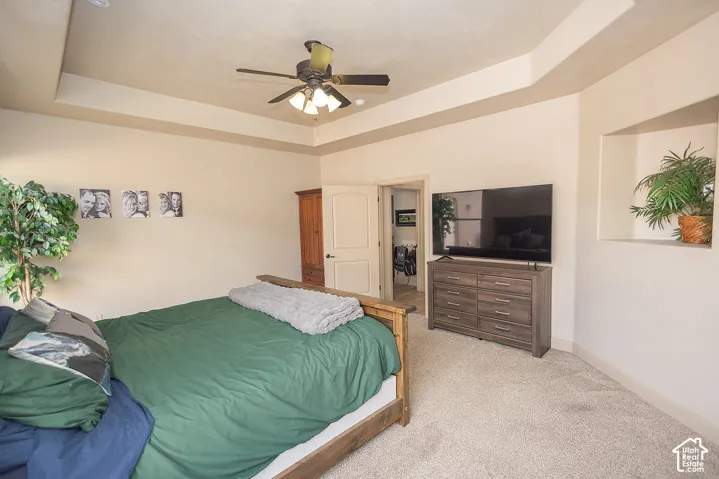 Tray ceiling with a ceiling fan light fixture, recessed wall niche, and light-toned carpet flooring