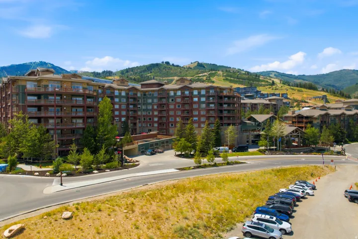 Multi-story building featuring multiple balconies, a structured parking area, and surrounding mature trees