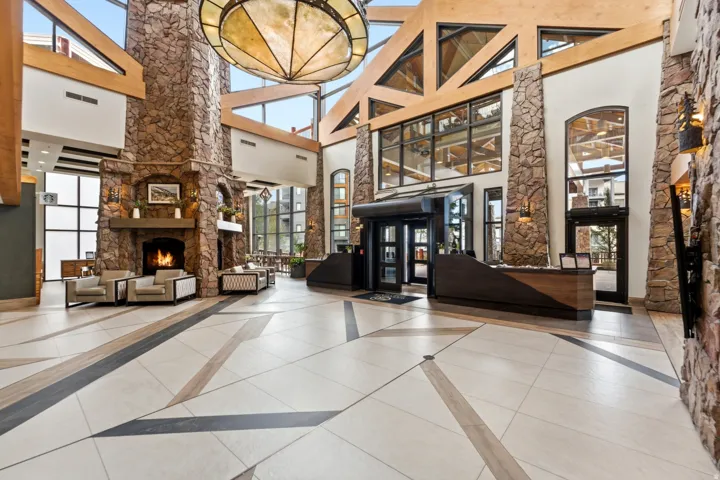 Vaulted ceiling with exposed wood beams, stone-clad columns, a stone fireplace with hearth, patterned tile flooring, and a reception desk