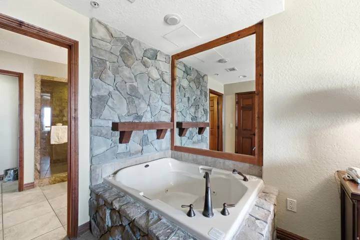 Built-in jetted tub surrounded by a stone-clad base, stone accent wall, large framed mirror, and warm wood trim