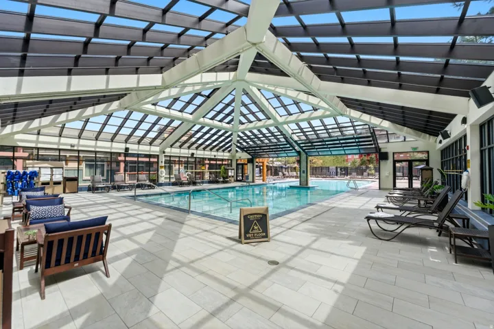Indoor swimming pool with a glass-paneled ceiling, light-toned tile flooring, and a glass wall opening to an outdoor pool area