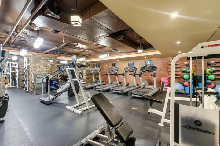 Dedicated fitness area featuring rubber flooring, exposed industrial-style ductwork, recessed lighting, and a stone-clad wall
