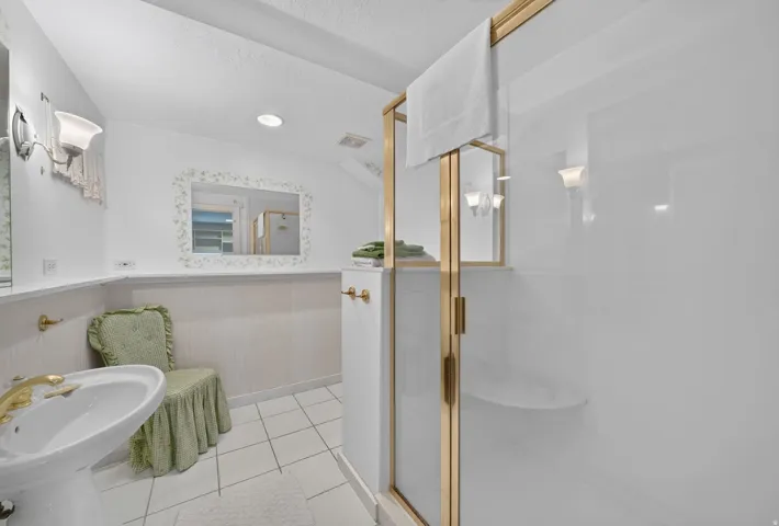 Pedestal sink with brass-finish faucet, vanity lighting, wall-mounted mirror, walk-in shower with gold-finish frame, and white tile flooring