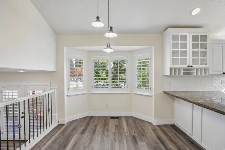 Bay window with plantation shutters, wood-finish flooring, white cabinetry with glass-front uppers, dark stone countertop, and pendant lighting