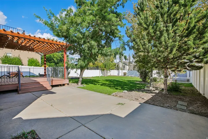 Expansive backyard featuring a wood-finish deck with a pergola, concrete patio space, and mature trees, all enclosed by a white privacy fence