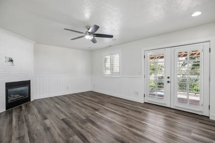 Wood-finish flooring, white wainscoting, white brick fireplace, ceiling fan, and French doors with plantation shutters