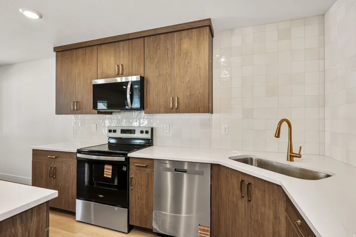 Modern kitchen featuring warm wood-finish cabinetry with brushed brass hardware, stainless steel appliances including a built-in microwave and dishwasher, white subway tile backsplash, and a waterfall-edge countertop with an undermount sink and brass fauc