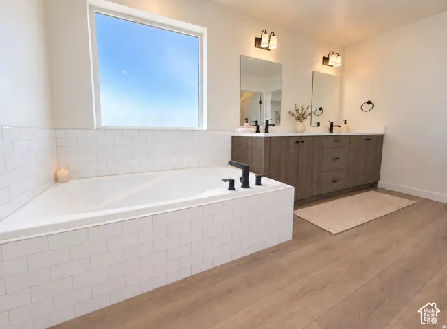 Corner soaking tub with subway tile surround, wood-finish flooring, dual vanity with undermount sinks, and matte black fixtures