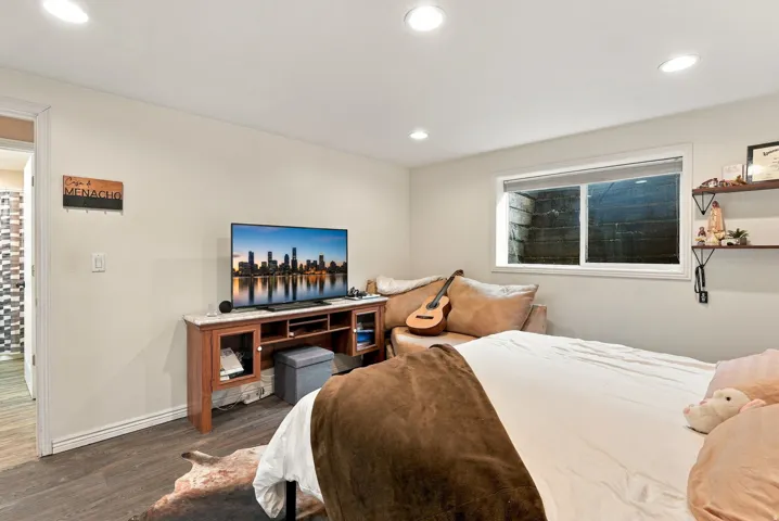 Recessed lighting illuminates the room, featuring wood-finish flooring and a window with a light-filtering shade