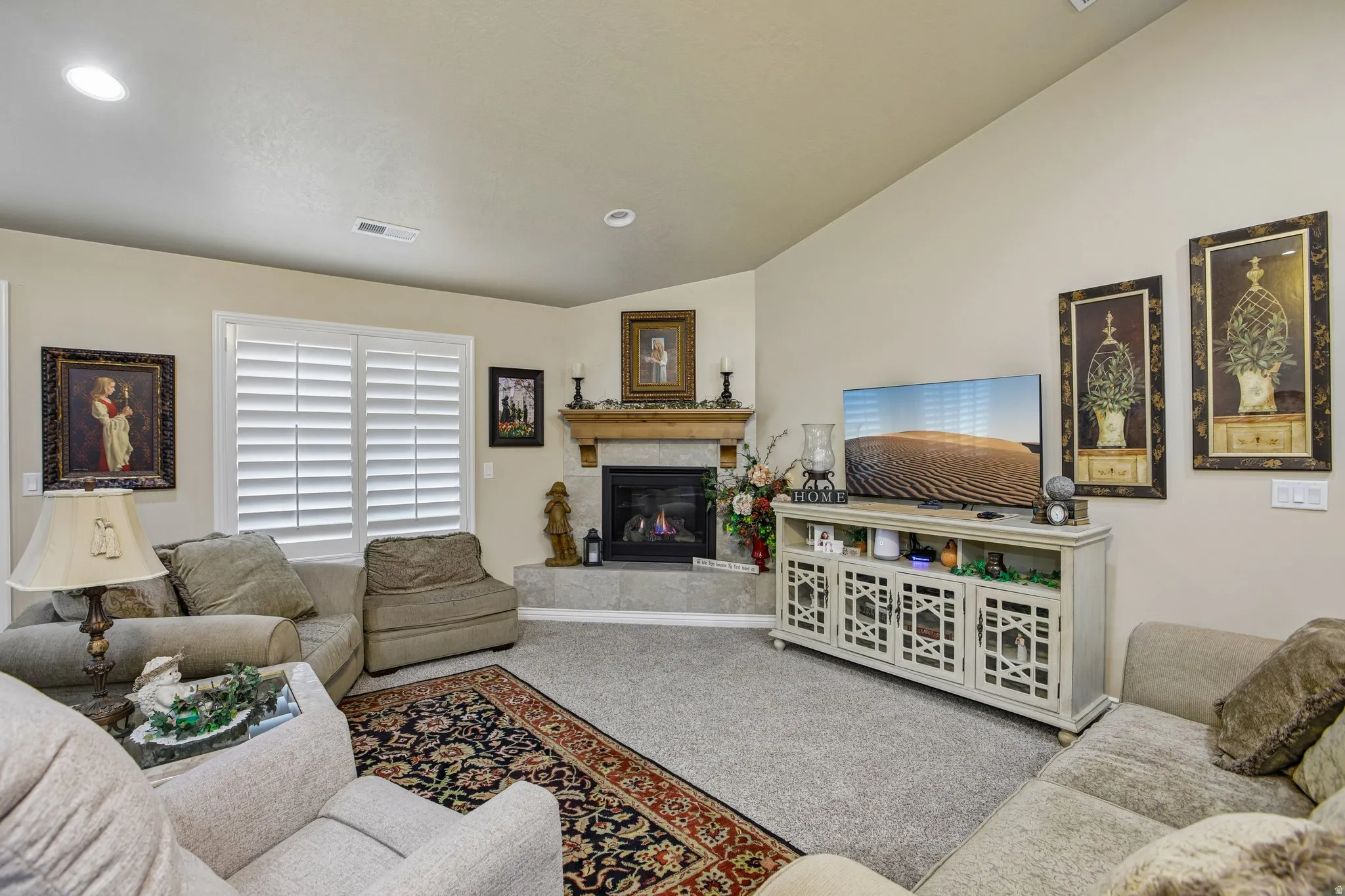 Vaulted ceiling, corner fireplace with stone surround, wood mantel, carpeted flooring, and window with plantation shutters