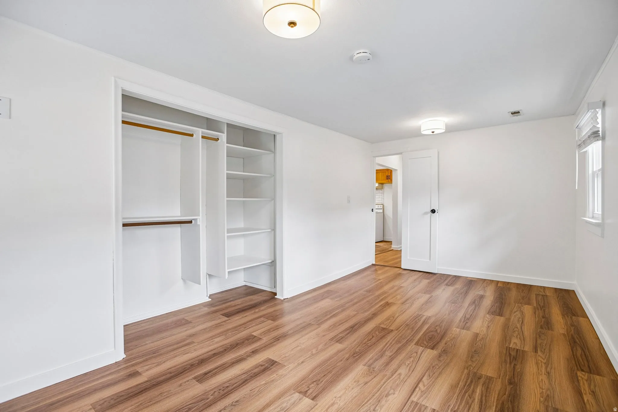 Wood-finish flooring, built-in closet organization, contemporary ceiling light fixture, white walls, and a window with blinds