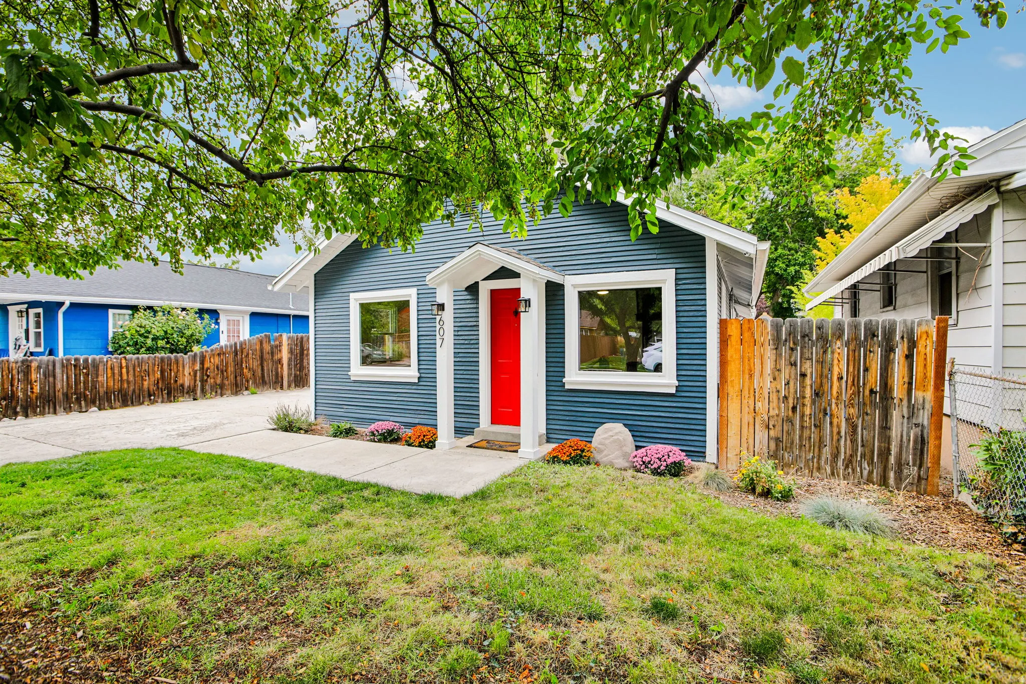 Blue corrugated siding, white trim windows, red front door, concrete walkway, and a landscaped garden bed