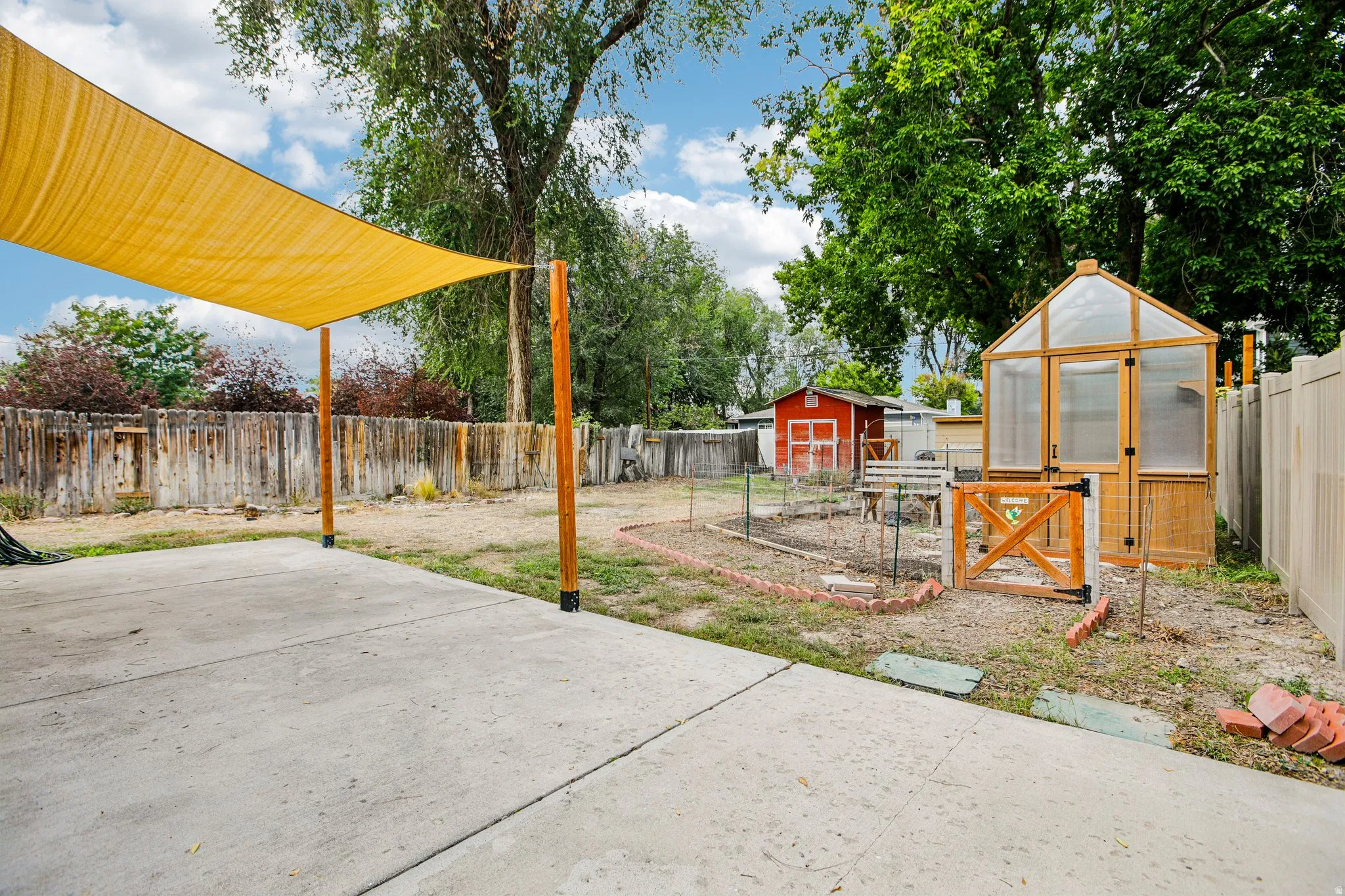 Concrete patio with an overhead shade sail, accompanied by a wooden-framed greenhouse, garden beds, and a red storage shed