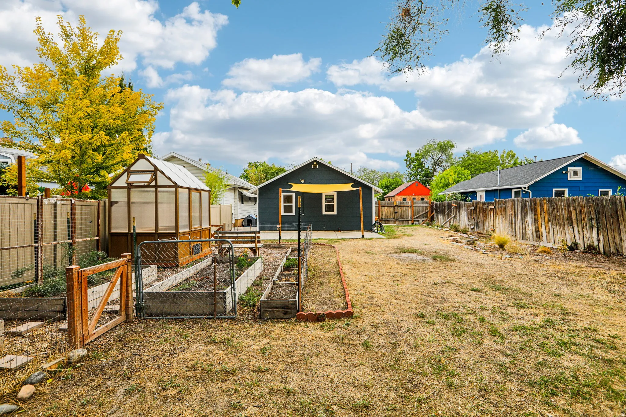 Spacious fenced yard featuring a greenhouse structure, multiple raised garden beds, and a back patio area with a sunshade