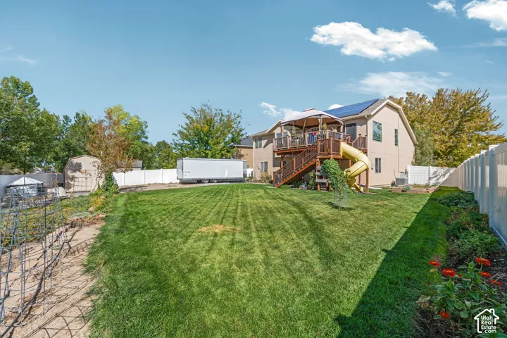Expansive backyard featuring a manicured lawn, elevated wood deck with stair access, integrated slide, solar panels, and perimeter privacy fencing