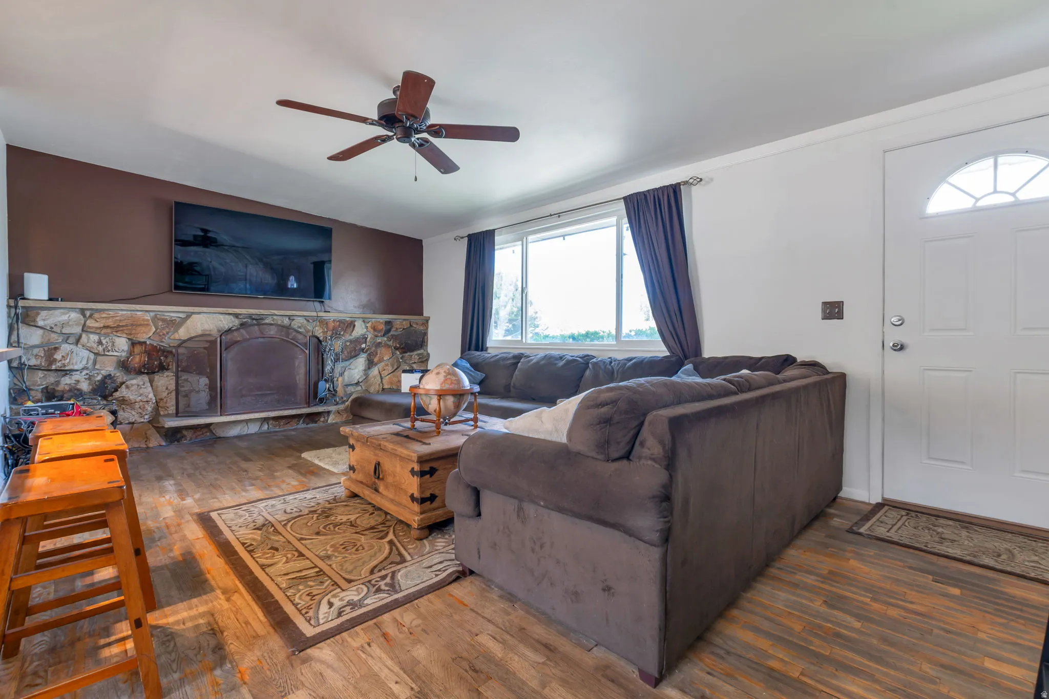 Spacious living area featuring a stone fireplace with hearth, wood-finish flooring, ceiling fan, and large windows