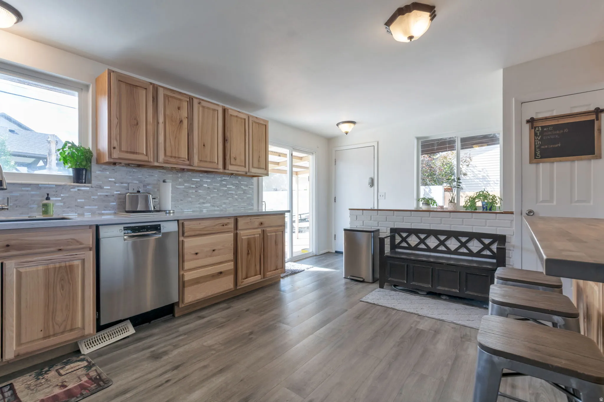Kitchen featuring light wood cabinetry, a mosaic tile backsplash, stainless steel dishwasher, light wood-finish flooring, and a sliding glass door