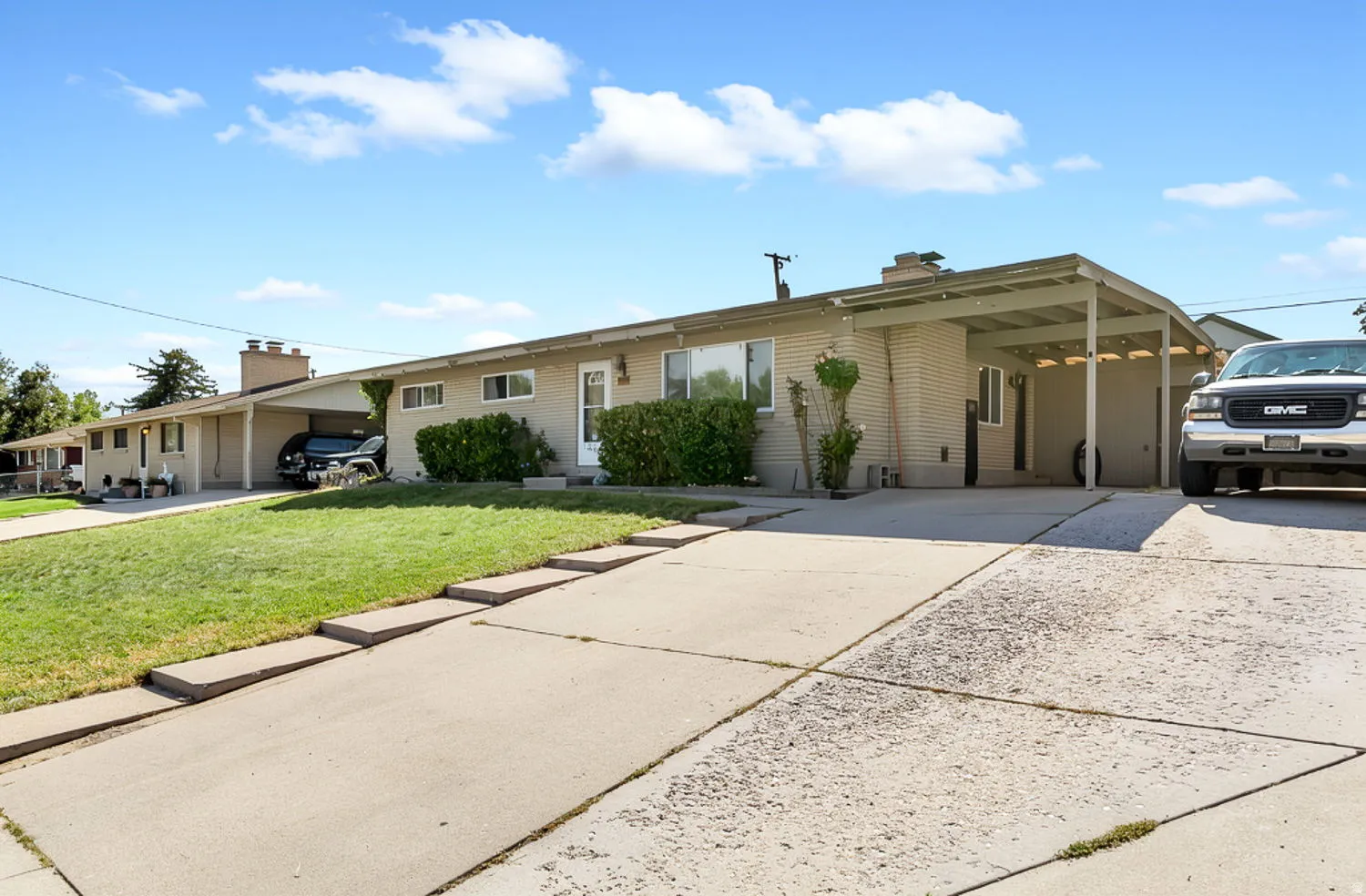 Light-colored brick exterior, covered carport, mature landscaping, concrete driveway, and front lawn